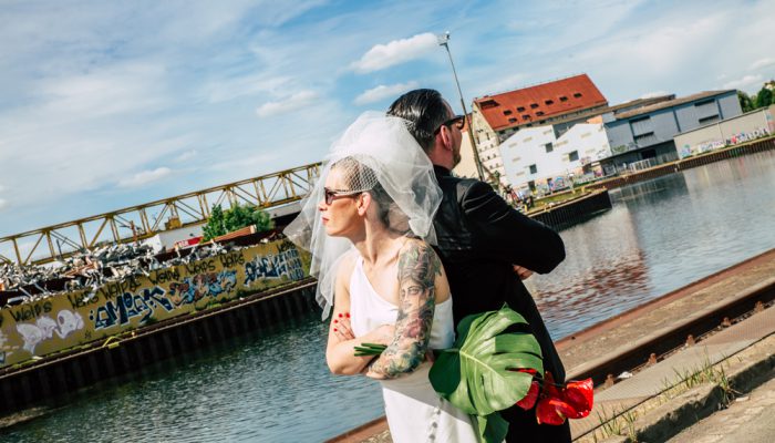 Brautpaar mit Sonnenbrillen posiert urban am Hafenbecken – Braut mit Tattoo und Monstera-Blume
