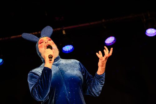 burlesque_02 Burlesque-Künstler Scotty Bunny steht im blauen Bunny-Kostüm auf der Bühne und singt unter farbigem Bühnenlicht.