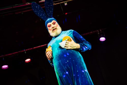burlesque_03 Scotty Bunny moderiert im blauen Bunny-Kostüm und Nipple Pasties expressiv vor dunklem Bühnenhintergrund.