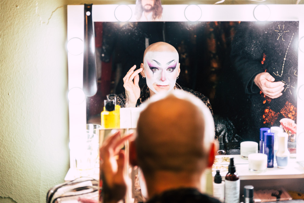 burlesque_035 Performer*in mit ausdrucksstarkem Bühnen-Make-up blickt konzentriert in den beleuchteten Spiegel während der Vorbereitung backstage.