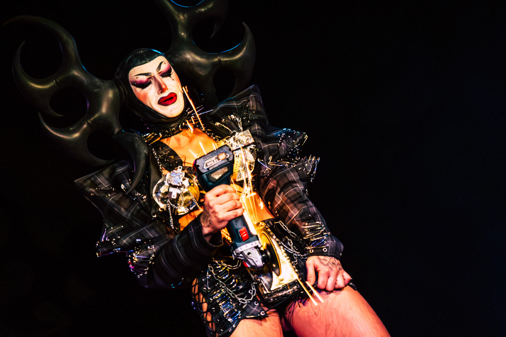 burlesque_04 Harpy Fatale performt auf der Bühne mit einer Schleifmaschine, Funken sprühen vor dunklem Hintergrund.