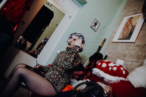 burlesque_047 Burlesque-Performer*in sitzt backstage im Kostüm auf einem Sofa und richtet Kopfschmuck und Make-up, umgeben von Requisiten und Garderobe.