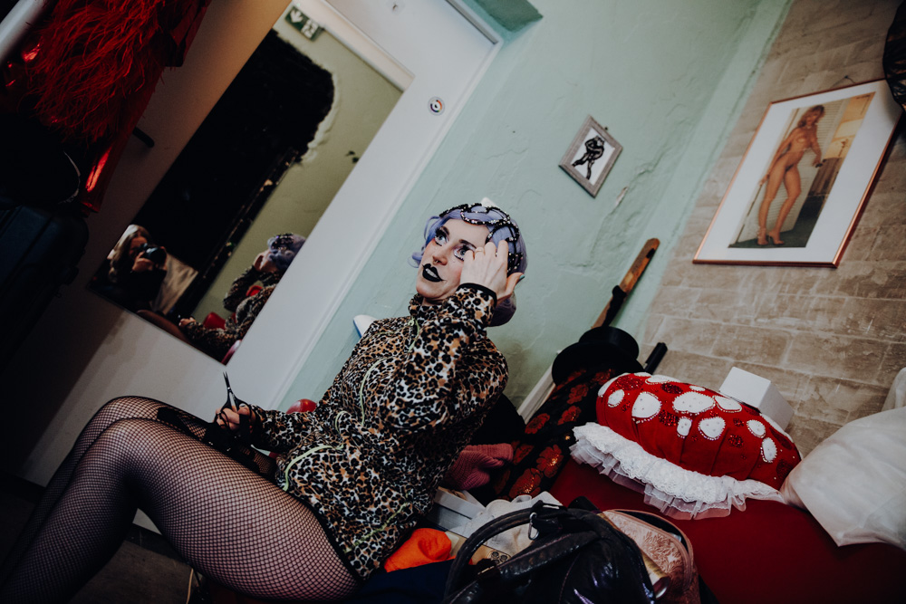 burlesque_047 Burlesque-Performer*in sitzt backstage im Kostüm auf einem Sofa und richtet Kopfschmuck und Make-up, umgeben von Requisiten und Garderobe.