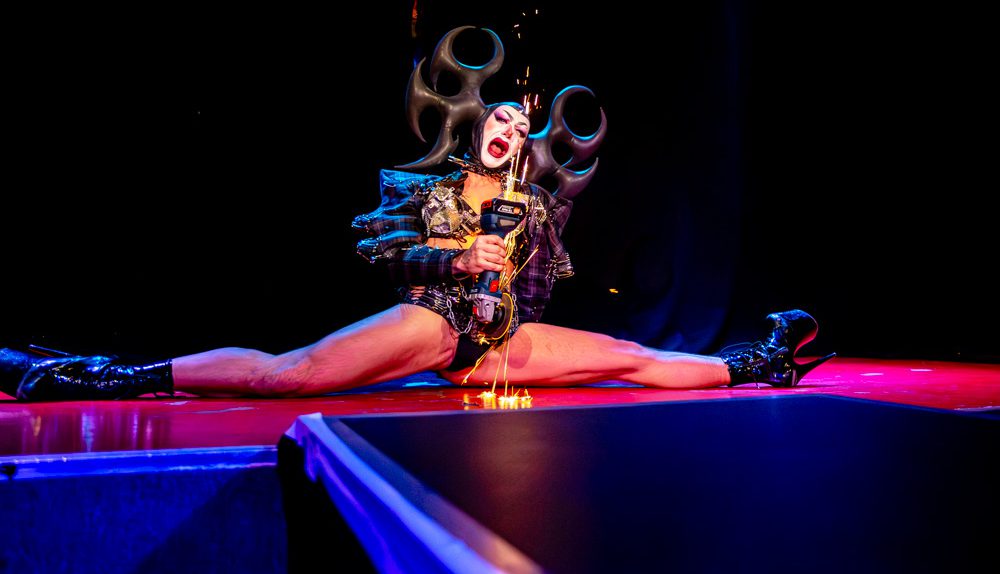 Harpy Fatale sitzt im Spagat auf der Bühne und performt mit einer Schleifmaschine, Funken sprühen über den Boden.