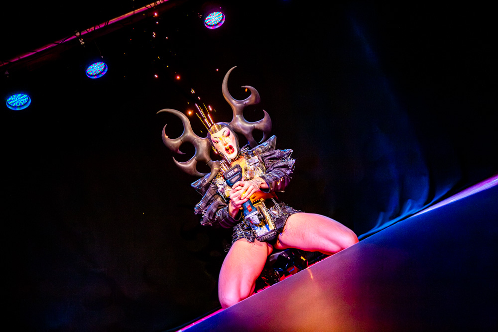 burlesque_07 Harpy Fatale steht kraftvoll auf der Bühne mit Schleifmaschine, Funken steigen auf unter farbigem Licht.