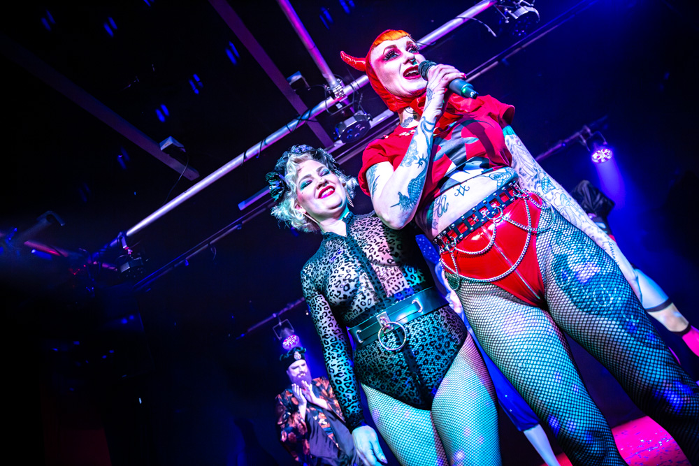 burlesque_19 Zwei Burlesque-Performerinnen stehen auf der Bühne, eine singt ins Mikrofon, beide im farbigen Bühnenlicht.