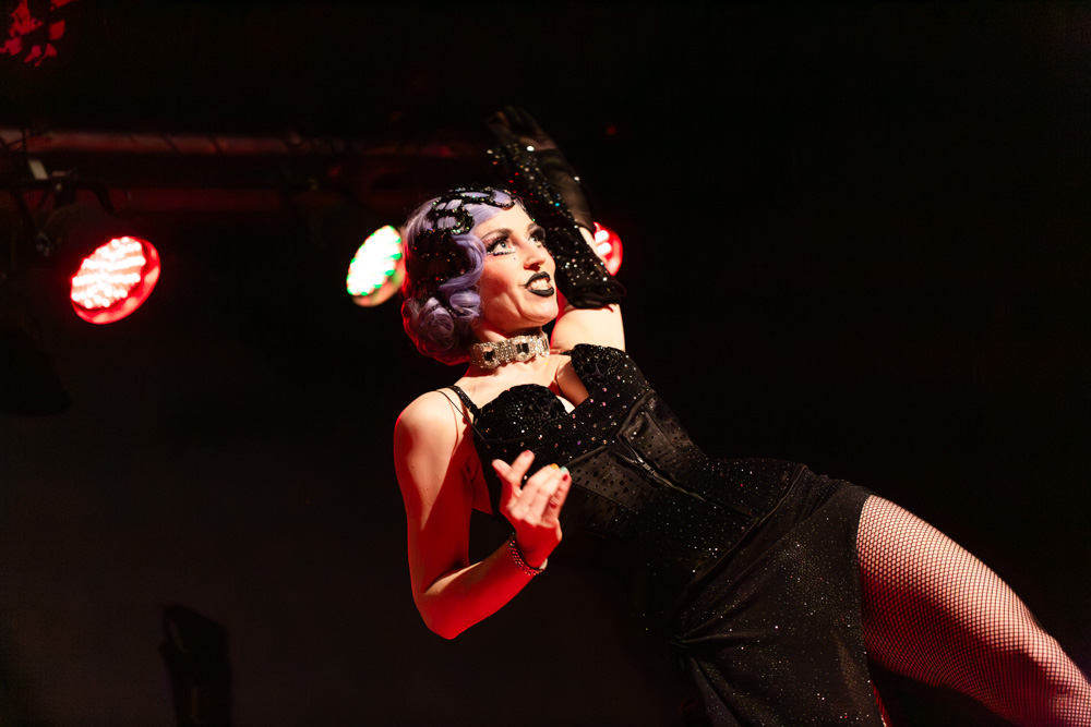 burlesque_23 Burlesque-Performer*in in schwarzem Bühnenkostüm mit Handschuhen und Netzstrümpfen, dynamische Bewegung unter rotem und grünem Bühnenlicht.