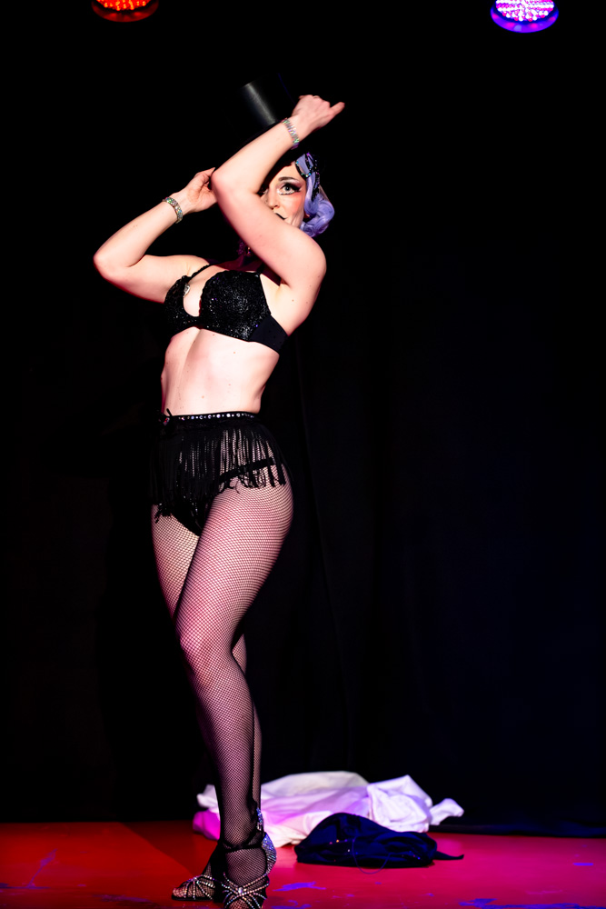 burlesque_24 Burlesque-Künstlerin Masha Champanskaya steht auf der Bühne in schwarzem Kostüm mit Netzstrümpfen und hebt die Arme in einer eleganten Pose unter farbigem Bühnenlicht.