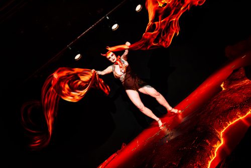 burlesque_31 Burlesque-Künstlerin Tronicat la Miez steht auf der Bühne und schwingt fließende rote Stoffbahnen in einer dynamischen Performance unter starkem Bühnenlicht.