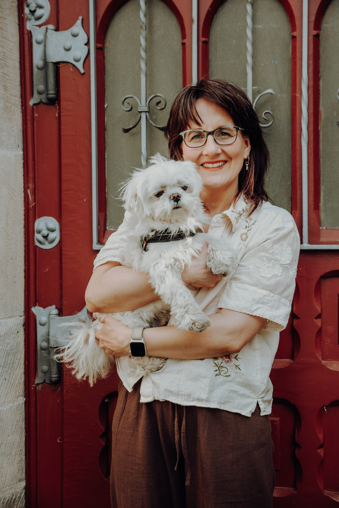 Maya Birken hält einen weißen Malteserhund vor einer roten historischen Haustür im Arm.