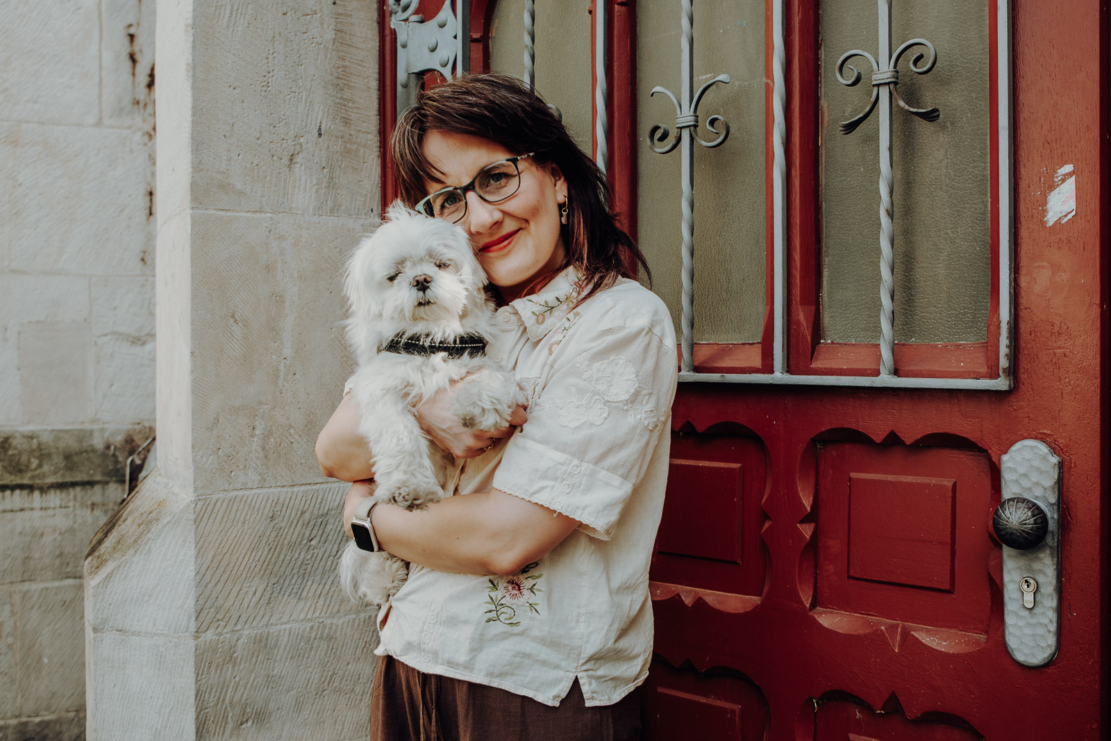 Frau mit Brille hält einen weißen Malteserhund vor einer roten historischen Haustür im Arm.