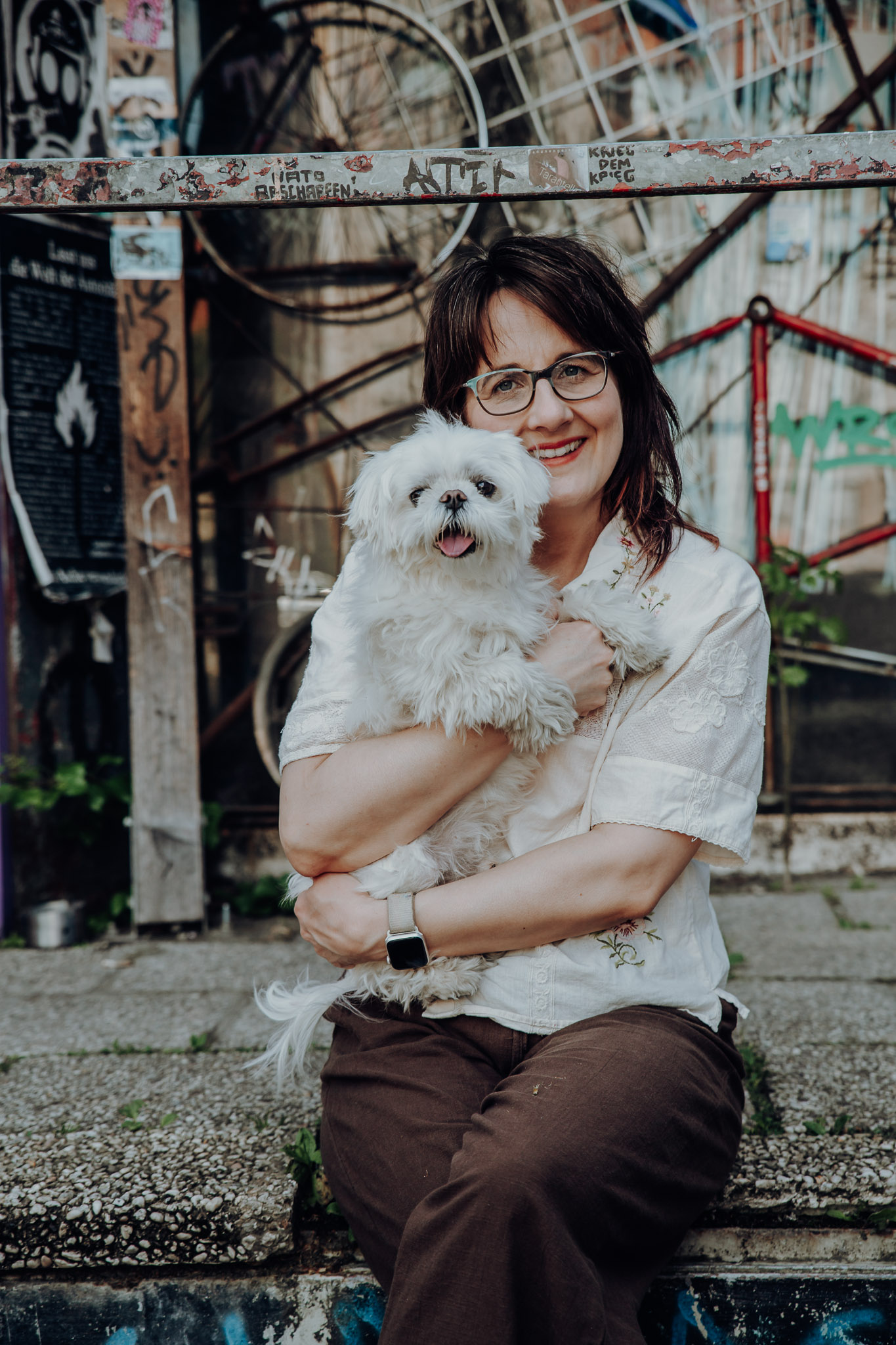 Frau mit Brille hält einen weißen Malteserhund bei einem urbanen Outdoor-Shooting im Arm.