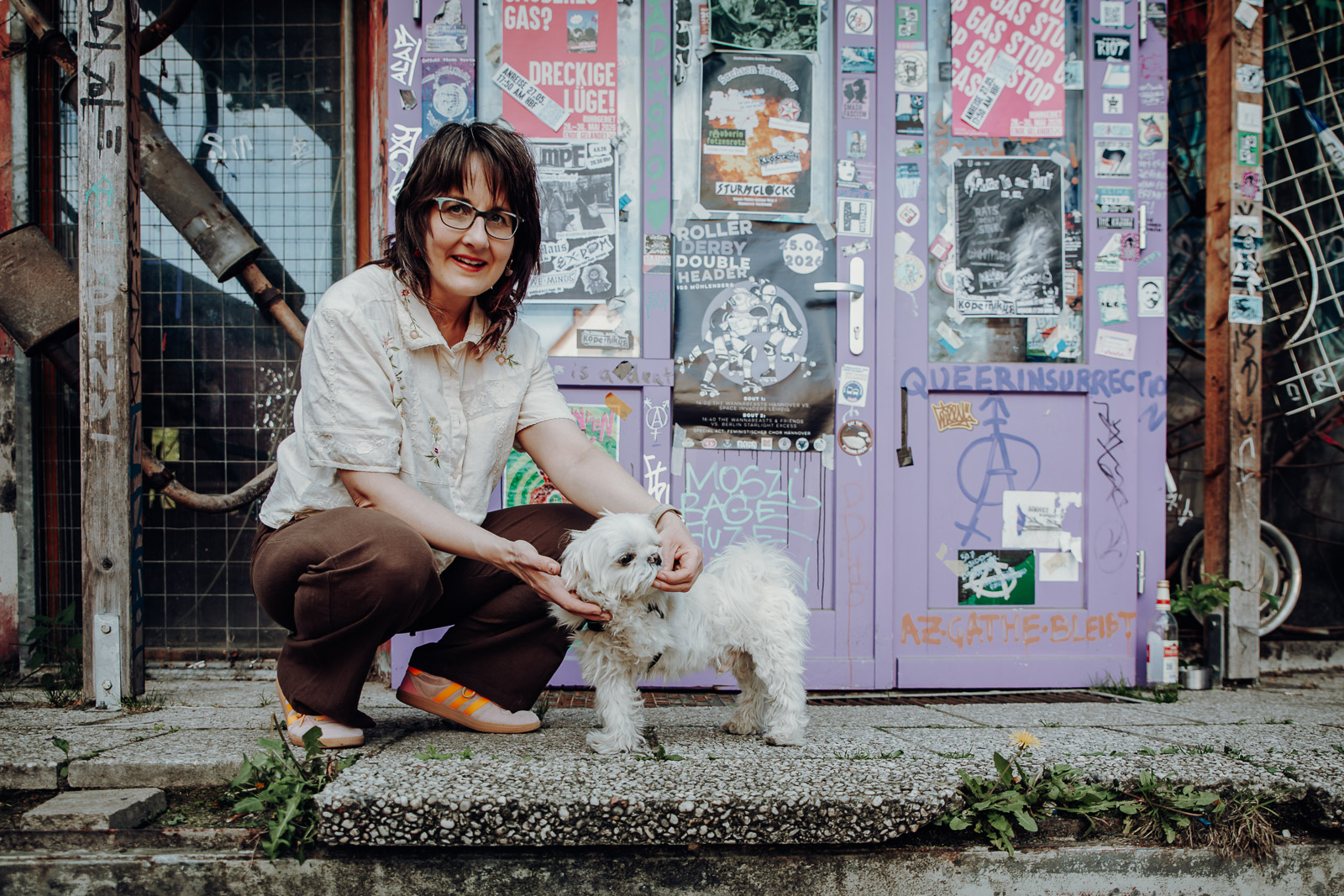 Frau kniet neben einem weißen Malteserhund vor einer mit Stickern und Plakaten bedeckten urbanen Kulisse.
